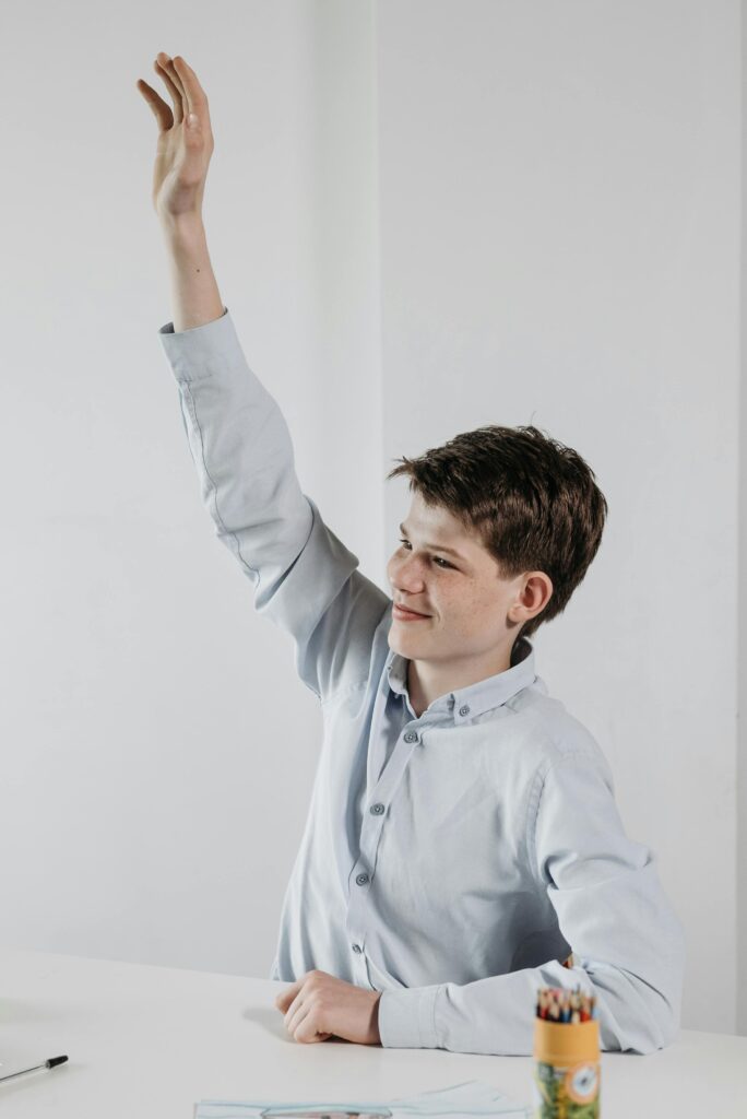 A smiling student in a light shirt raising hand at a desk indoors.